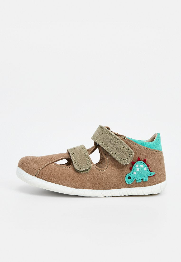 Brauner Kleinkindschuh mit zwei Klettverschlüssen, weißer Sohle, türkisfarbenem Innenfutter und einem kleinen blauen Dinosaurier-Aufnäher an der Seite.