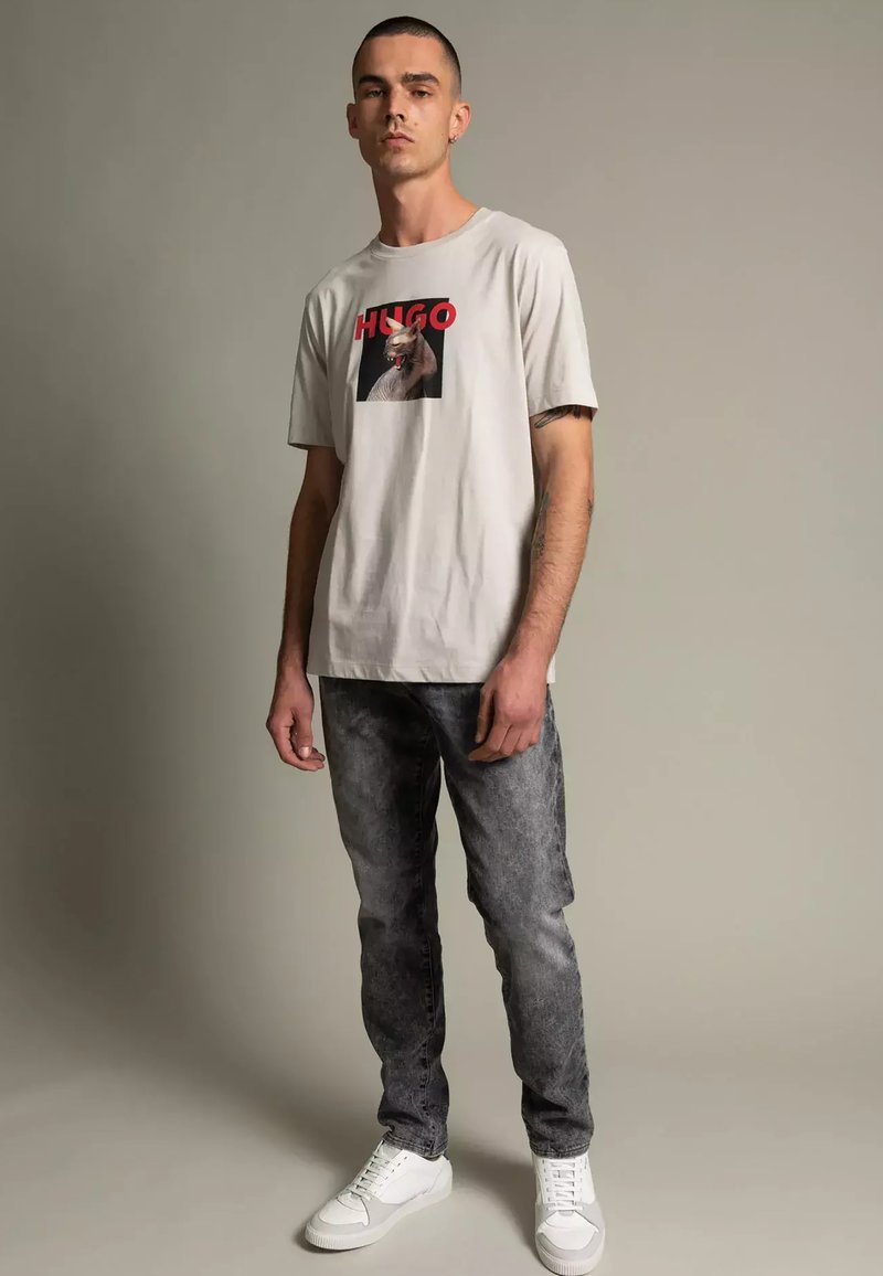 Hellgraues T-Shirt mit einem grafischen Druck mit dem Text "HUGO". Kombiniert mit dunkelgrauen, ausgewaschenen Jeans und weißen Sneakern.