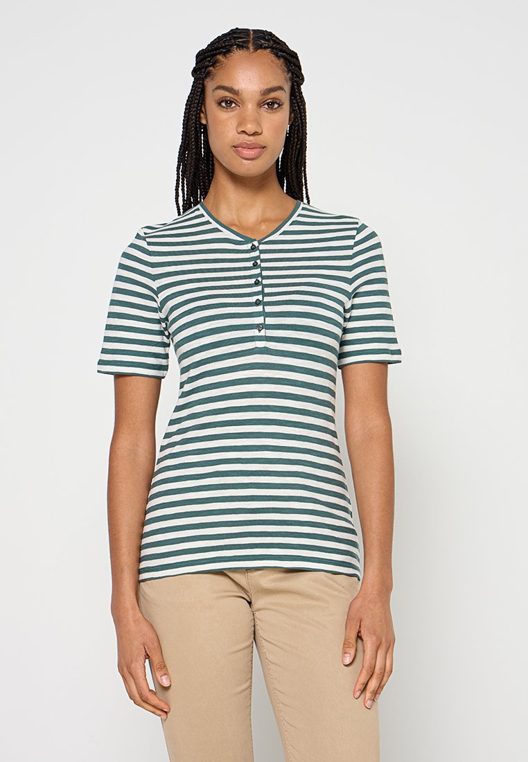 Frau trägt ein grün-weiß gestreiftes Henley-Shirt mit kurzen Ärmeln und beige Hosen, steht vor einem schlichten weißen Hintergrund.