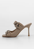 Beige High-Heel-Mule-Sandale mit zwei breiten Riemen, großen silbernen Schnallen und einem schlanken Stiletto-Absatz auf einem einfarbigen Hintergrund.