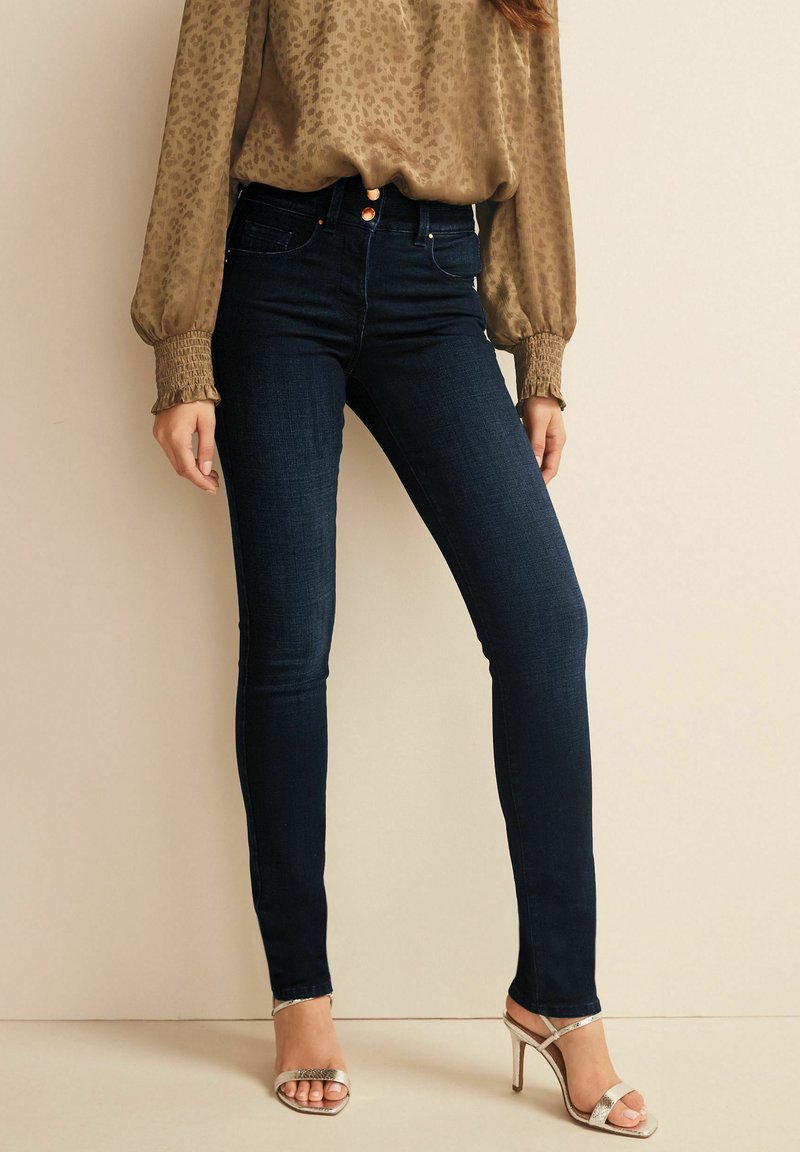 Frau trägt dunkelblaue Skinny Jeans, eine hellbraune Bluse mit langen Ärmeln und gerafften Manschetten sowie silberne High-Heels.