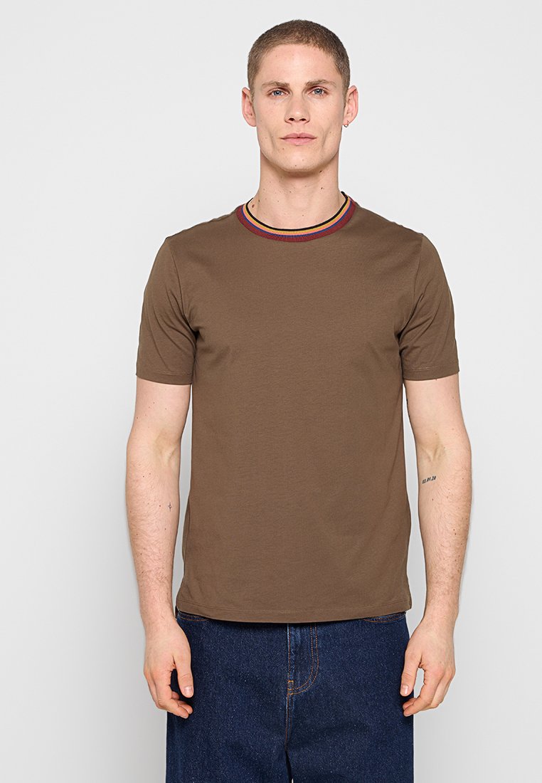 Mann trägt braunes T-Shirt mit kurzen Ärmeln und gestreiftem Kragen sowie blaue Jeanshose, steht vor einem einfarbigen hellgrauen Hintergrund.
