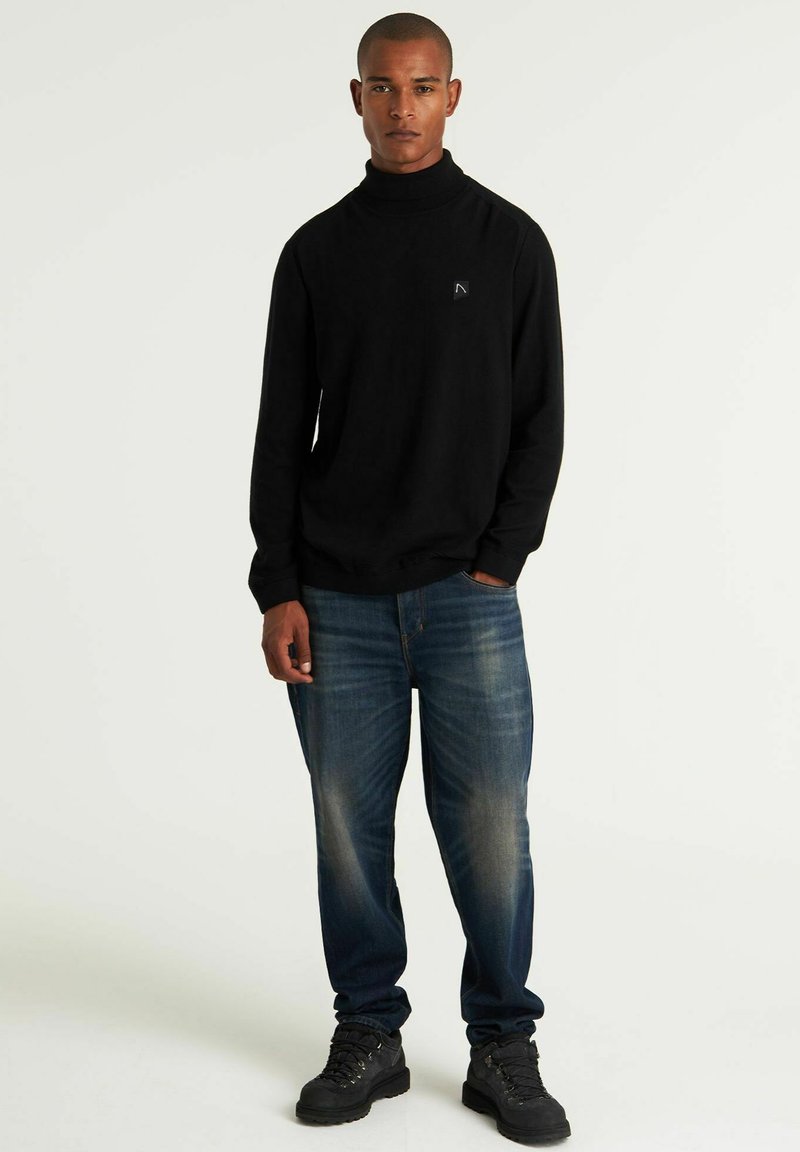Schwarzer Rollkragenpullover aus weichem Material, kombiniert mit blauen Jeans aus Denim mit einem ausgewaschenen Muster. Das Modell trägt schwarze Wanderschuhe.