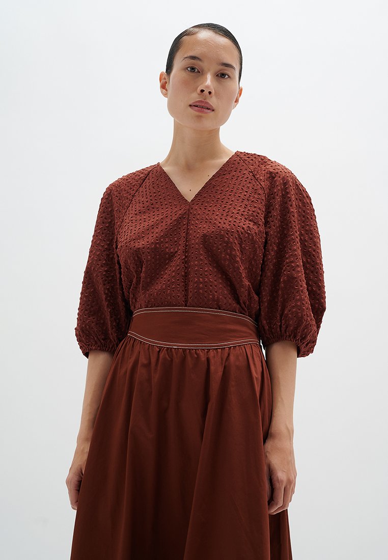 Rostfarbige Bluse mit strukturiertem Muster, puffartigen Ärmeln und V-Ausschnitt. Kombiniert mit einem passenden Rock mit Taillenbund, was ein aufeinander abgestimmtes Outfit ergibt.