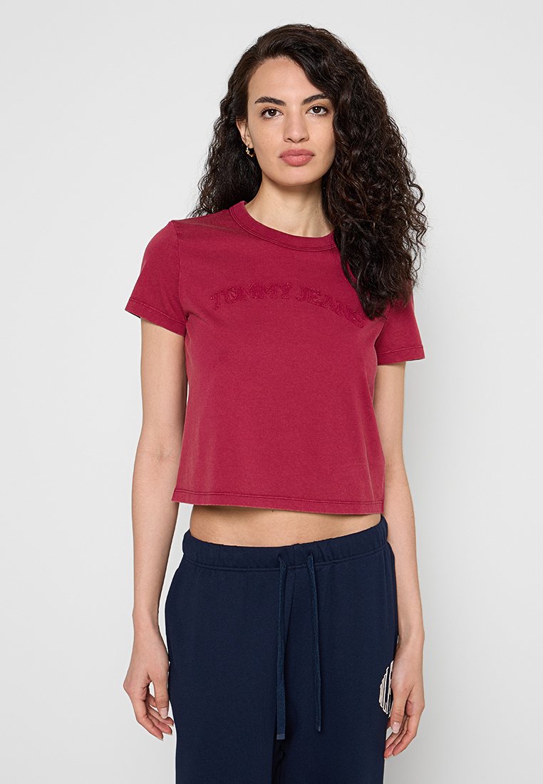 Junge Frau mit lockigem Haar, die ein rotes, kurzes Tommy Jeans T-Shirt und marineblaue Jogginghosen trägt, steht vor einem schlichten Hintergrund.