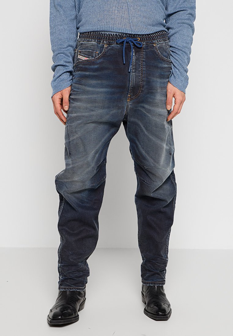 Mann trägt ausgewaschene dunkelblaue Denim-Jogginghosen kombiniert mit schwarzen Schnürschuhen und einem hellblauen, gerippten Langarmshirt.