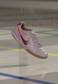 Hellvioletter Nike-Hallenschuh für Fußball mit dunkelviolettem Swoosh und pinker Umrandung, schwebt über einem Hallenboden mit farbigen Linien.