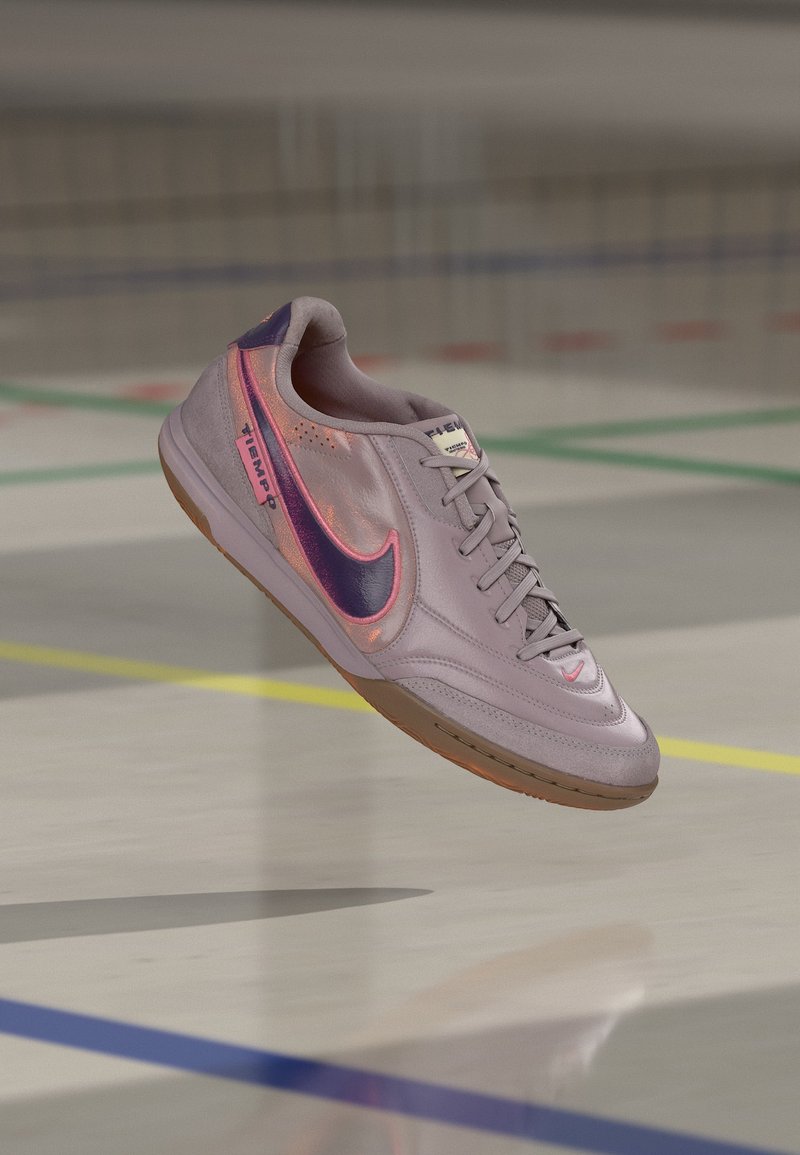 Hellvioletter Nike-Hallenschuh für Fußball mit dunkelviolettem Swoosh und pinker Umrandung, schwebt über einem Hallenboden mit farbigen Linien.