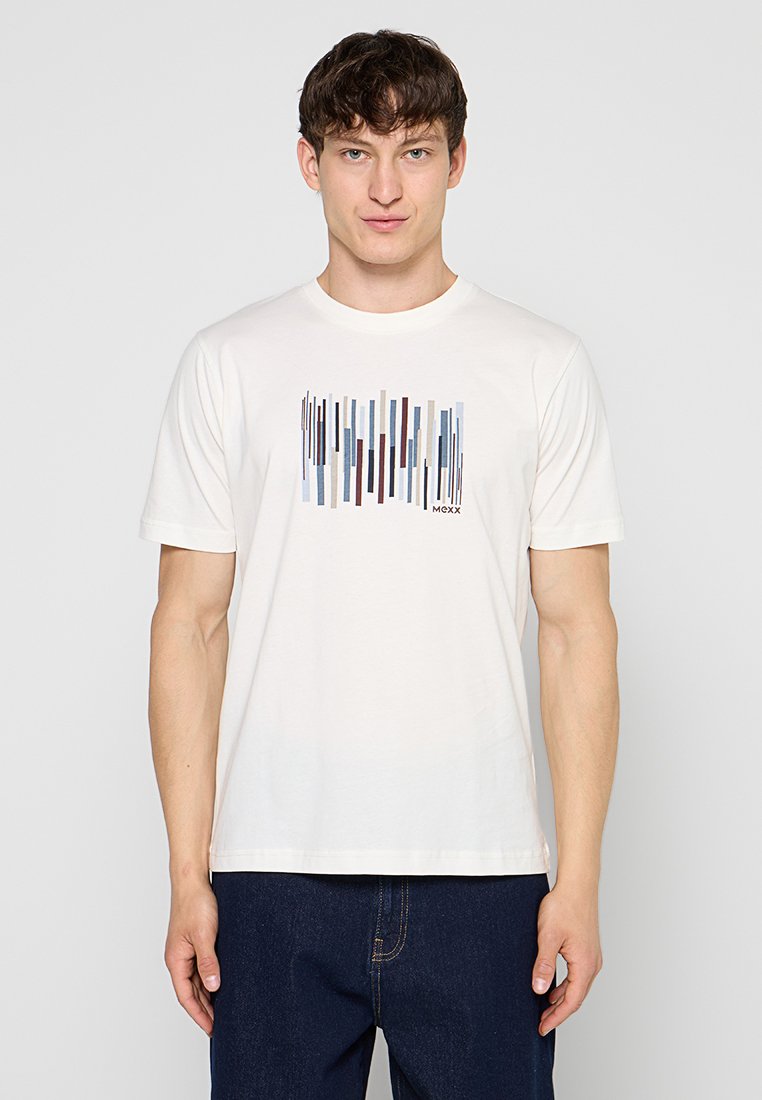Junger Mann, der ein weißes T-Shirt mit einem mehrfarbigen vertikalen Streifenmuster und dunkelblaue Jeans trägt, steht vor einem schlichten hellen Hintergrund.