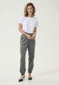 Weißes T-Shirt, graue taillierte Hose mit elastischen Bündchen und schwarze Absatzschuhe. Lässiges Outfit mit einfachen Linien und minimalem Schmuck.