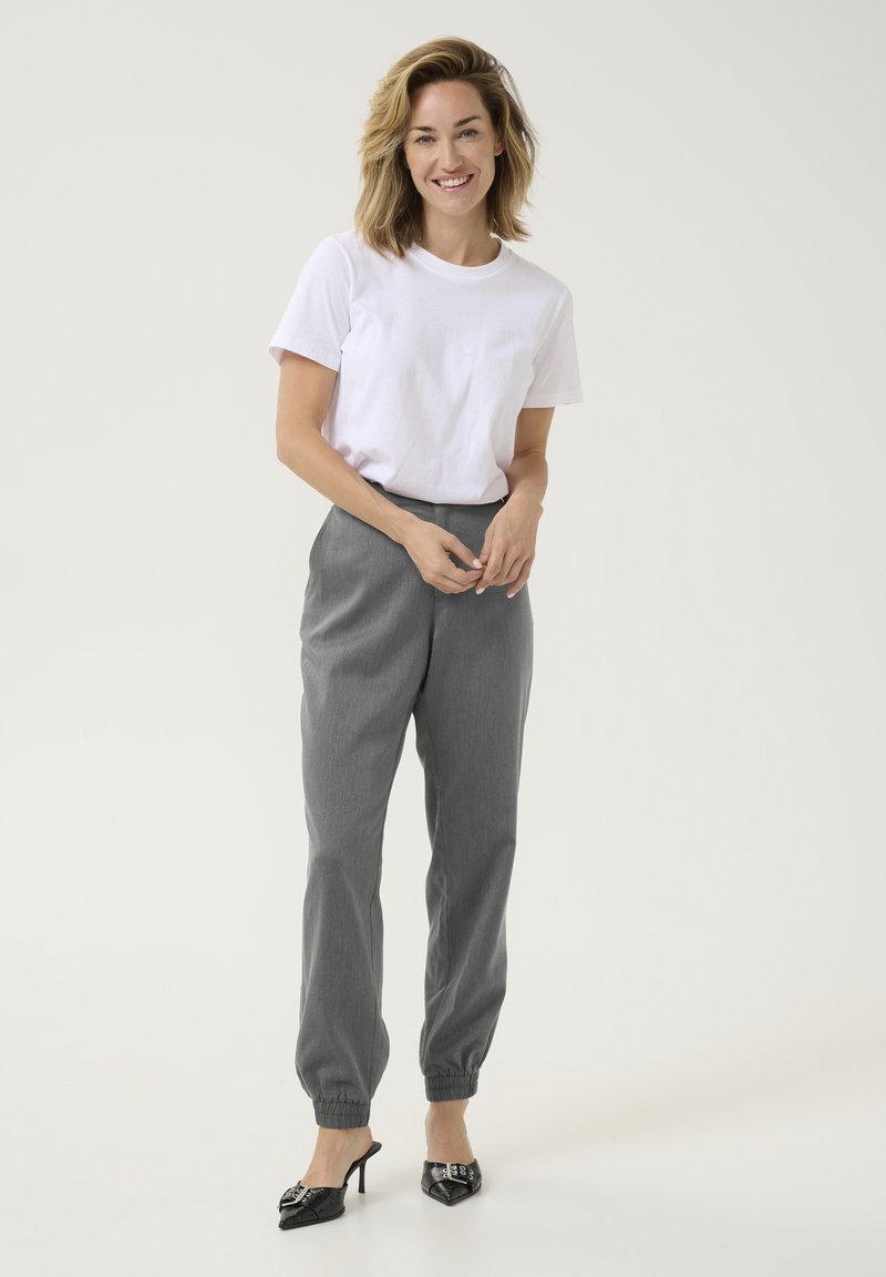 Weißes T-Shirt, graue taillierte Hose mit elastischen Bündchen und schwarze Absatzschuhe. Lässiges Outfit mit einfachen Linien und minimalem Schmuck.