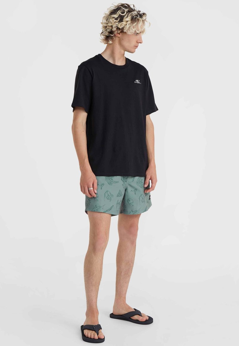 Schwarzes Baumwoll-T-Shirt kombiniert mit grünen, gemusterten Badeshorts mit Illustrationen. Das Model trägt schwarze Flip-Flops und steht seitlich.