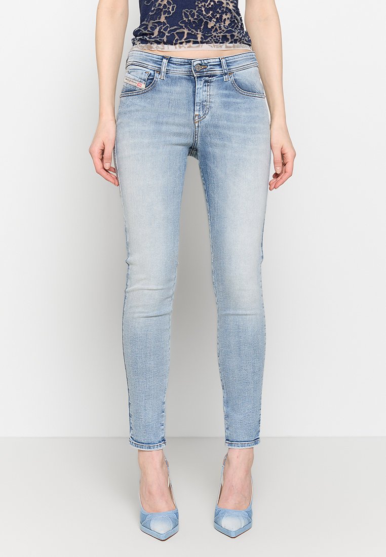 Frau in hellblauen Skinny Jeans, einem dunklen Spitzenoberteil mit floralen Muster und hellblauen spitzen High Heels, steht vor einem schlichten weißen Hintergrund.