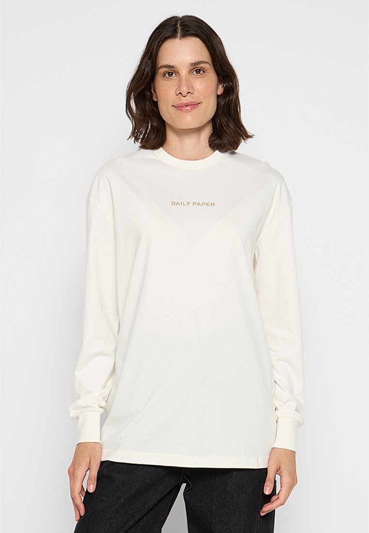 Frau mit schulterlangem dunklem Haar, die ein weißes langärmliges Shirt mit dem Text "DAILY PAPER" und schwarze Hosen trägt, steht vor einem schlichten Hintergrund.