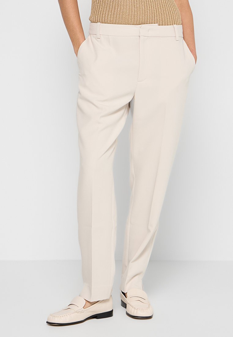 Beige maßgeschneiderte Hose mit geradem Schnitt, vorderen Taschen und glatter Oberfläche, kombiniert mit hellen Loafers.