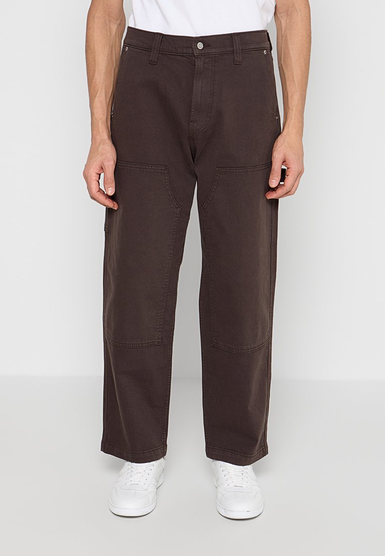 Mann trägt lockere, dunkelbraune Hose mit großen Vordertaschen und weiße Turnschuhe, steht vor einem einfachen, hellen Hintergrund.