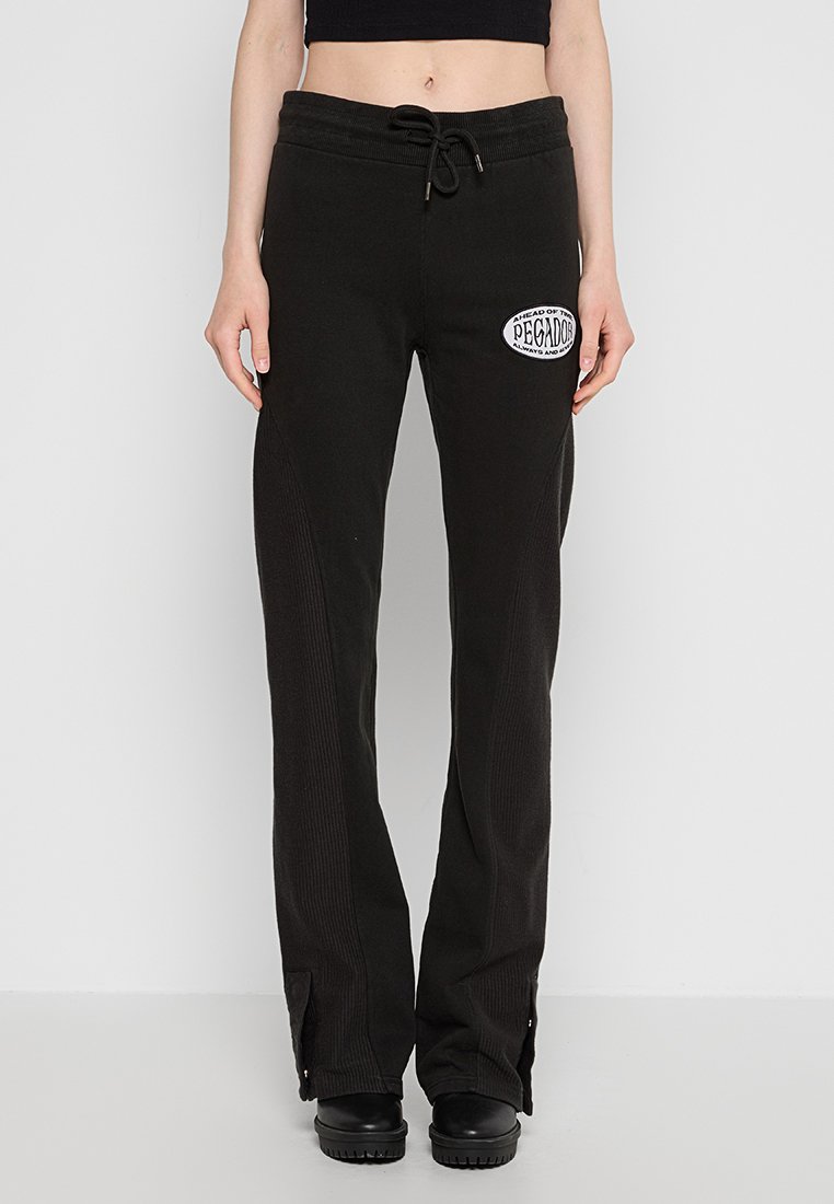 Schwarze Sweatpants mit strukturiertem Rippenmaterial, mit einem Kordelzugbund und einem weißen Patch mit Text am linken Oberschenkel. Eng an den Knöcheln.