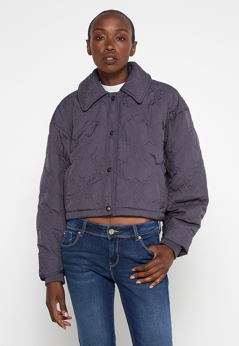 Gekürzte, dunkelgraue gesteppte Jacke mit großem Kragen und Knopfleiste, die über Rautenmuster und eine glatte Textur verfügt. Tragen mit blauen Jeans.