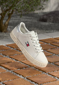 Weiße Sneaker aus synthetischem Material mit glatter Oberfläche, einer strukturierten Sohle und geprägtem "TOMMY JEANS"-Schriftzug. Silberner Akzent an der Ferse.