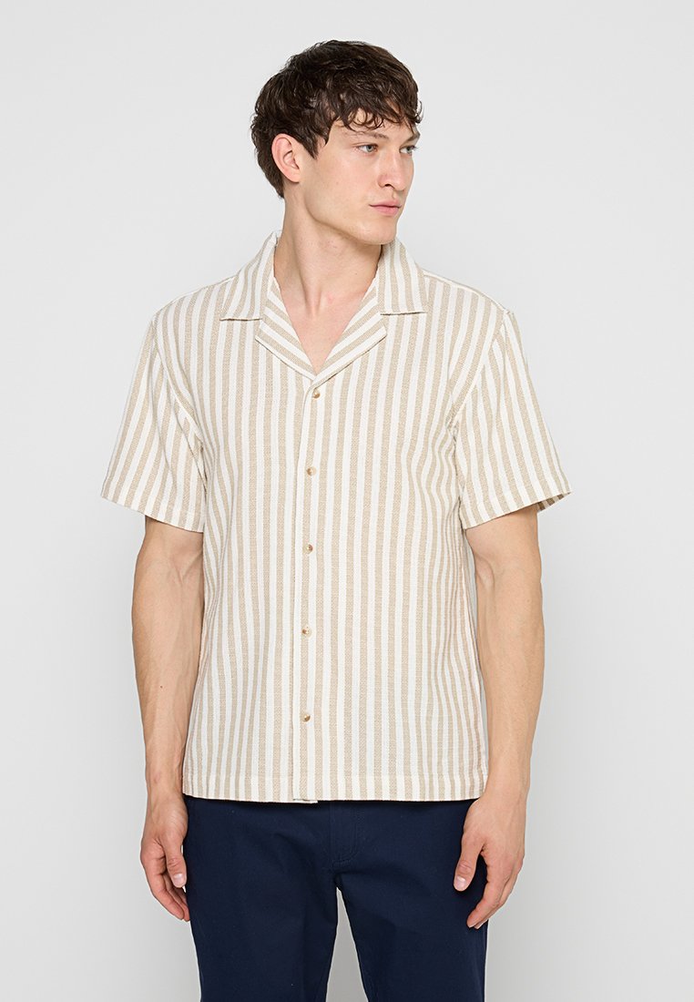 Junger Mann trägt ein beige-weiß vertikal gestreiftes kurzärmeliges Hemd mit Knöpfen und dunkelblaue Hose, schaut zur Seite.