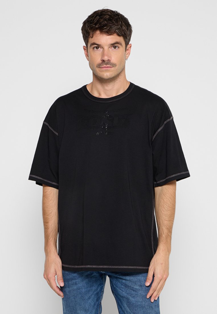 Schwarzes T-Shirt mit kurzen Ärmeln und Rundhalsausschnitt, mit tonalem Nähte und einem strukturierten Logo auf der Vorderseite. Getragen mit hellblauen Jeans.
