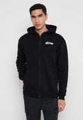 Junger Mann trägt einen schwarzen Zip-Hoodie mit dem "elrow"-Logo auf der Brust und steht mit einer Hand in der Tasche vor einem einfarbigen Hintergrund.