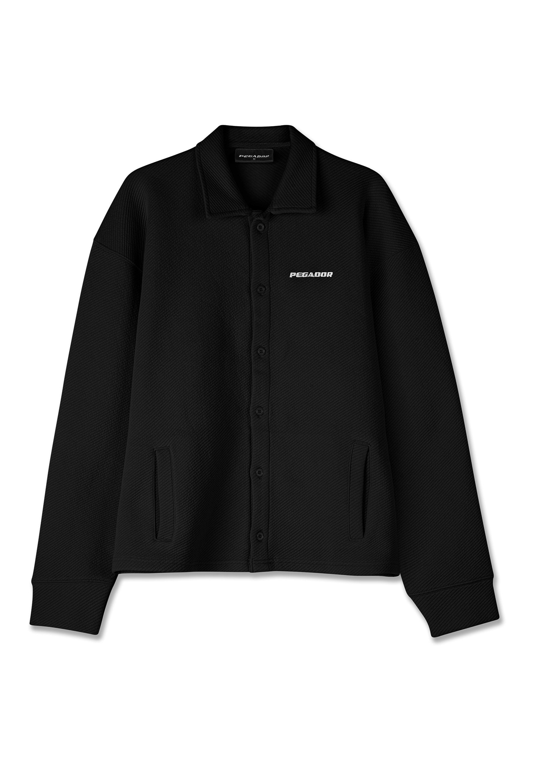 Schwarze Hemdjacke aus strukturiertem Stoff, mit Kragen, langen Ärmeln, seitlichen Taschen und einem Logo auf der linken Seite.
