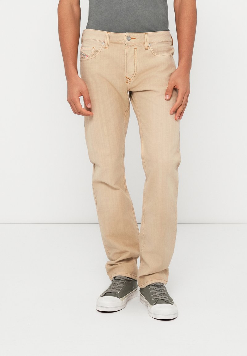 Hellbeige Jeans mit geradem Bein, subtiler Textur und orangefarbenen Zierstichen, getragen mit grünen Sneakern vor einfarbigem Hintergrund.
