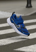 Blauer Sportschuh mit Mesh-Obermaterial, Gummisohle, verstellbarem Riemen und schwarzen sowie weißen Akzenten. Über einem Fußgängerüberweg platziert.