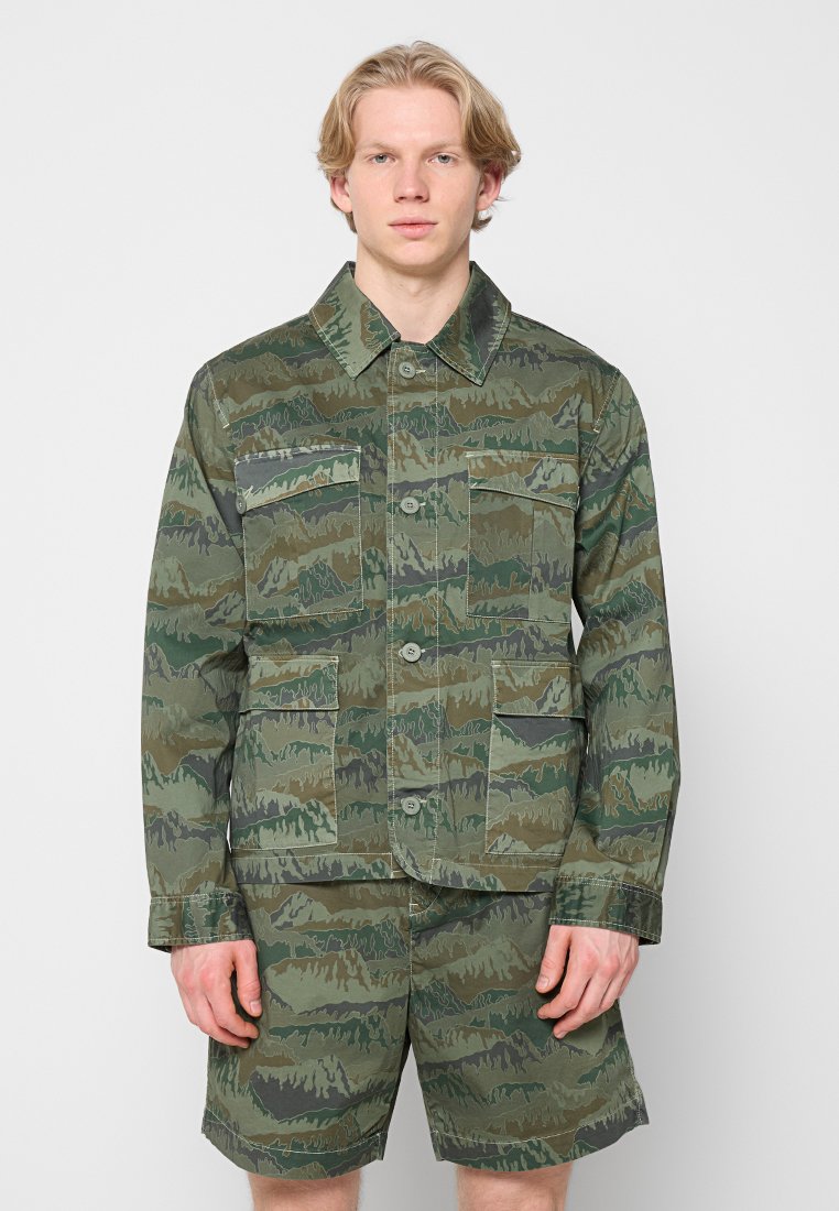 Jacke und Shorts im Tarnmuster in grünen und braunen Tönen. Verfügt über vier Vordertaschen und einen Knopfverschluss. Aus Baumwollmaterial.