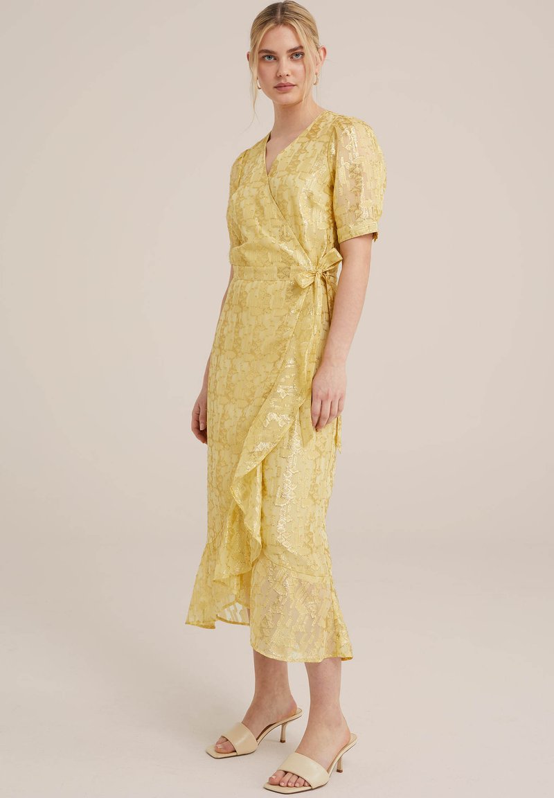 Wickelkleid aus gelber Spitze mit kurzen Puffärmeln, gerafftem Saum und Bindeband in der Taille. Kombiniert mit beigen Sandalen mit offenen Zehen.