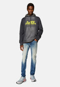 Grauer Pullover mit neonfarbigem gelbem Logo, hellblaue, ausgebleichte Jeans und graue Sneakers mit roten Akzenten. Das Model steht selbstbewusst mit den Händen in den Taschen.