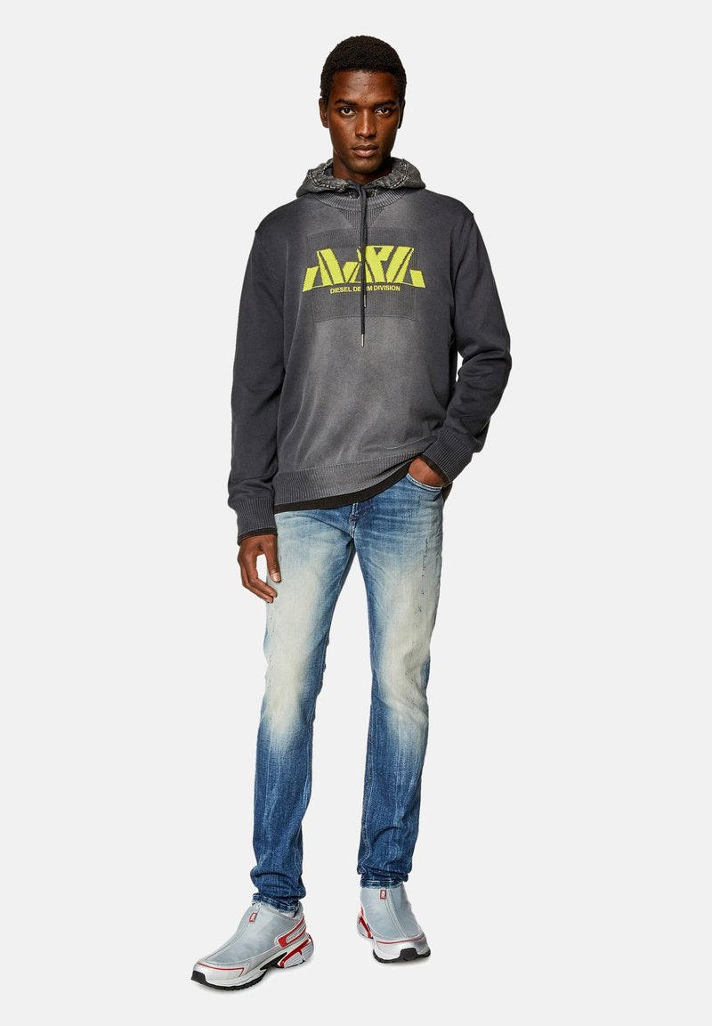 Grauer Pullover mit neonfarbigem gelbem Logo, hellblaue, ausgebleichte Jeans und graue Sneakers mit roten Akzenten. Das Model steht selbstbewusst mit den Händen in den Taschen.