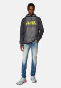Grauer Pullover mit neonfarbigem gelbem Logo, hellblaue, ausgebleichte Jeans und graue Sneakers mit roten Akzenten. Das Model steht selbstbewusst mit den Händen in den Taschen.
