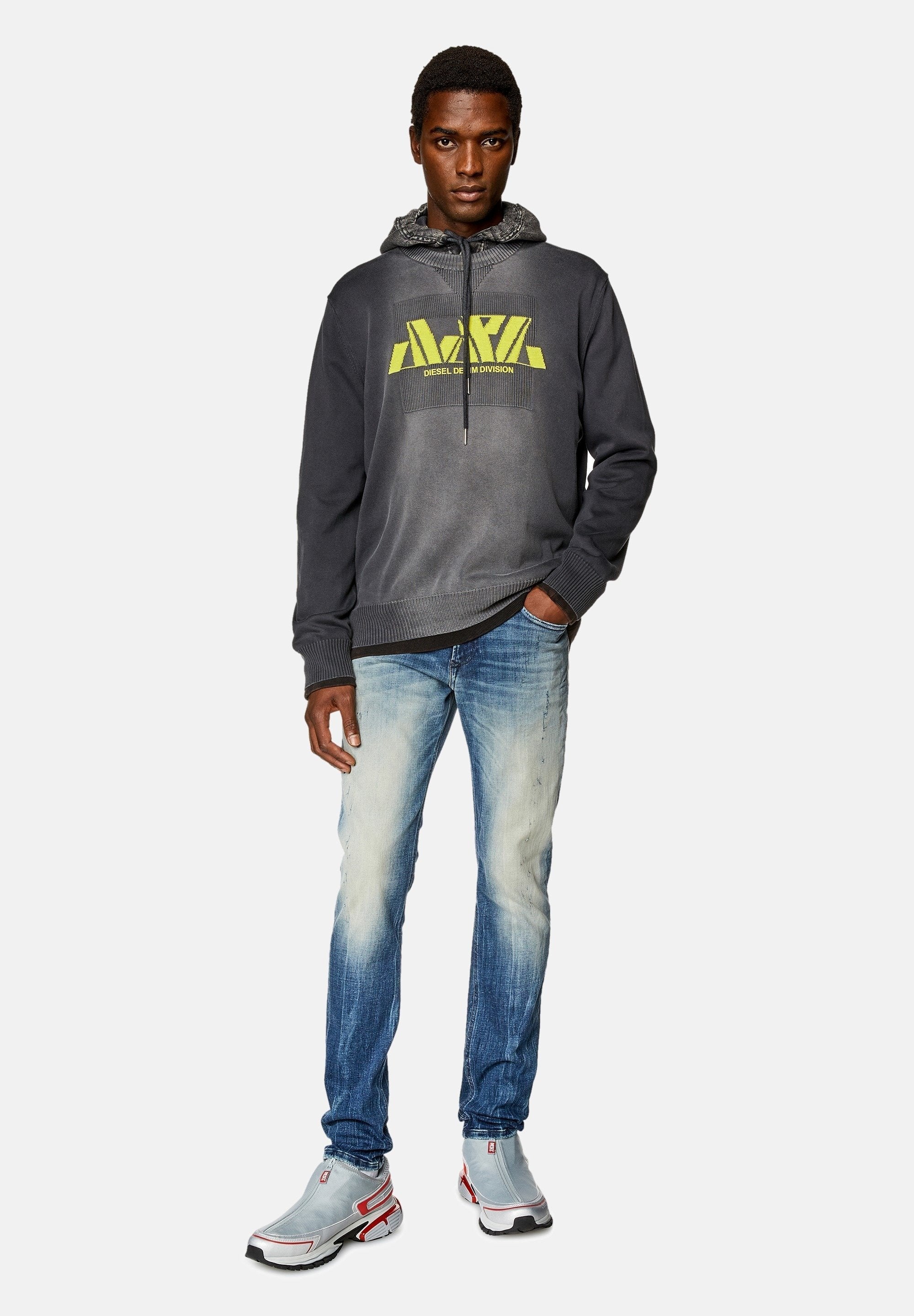 Grauer Pullover mit neonfarbigem gelbem Logo, hellblaue, ausgebleichte Jeans und graue Sneakers mit roten Akzenten. Das Model steht selbstbewusst mit den Händen in den Taschen.