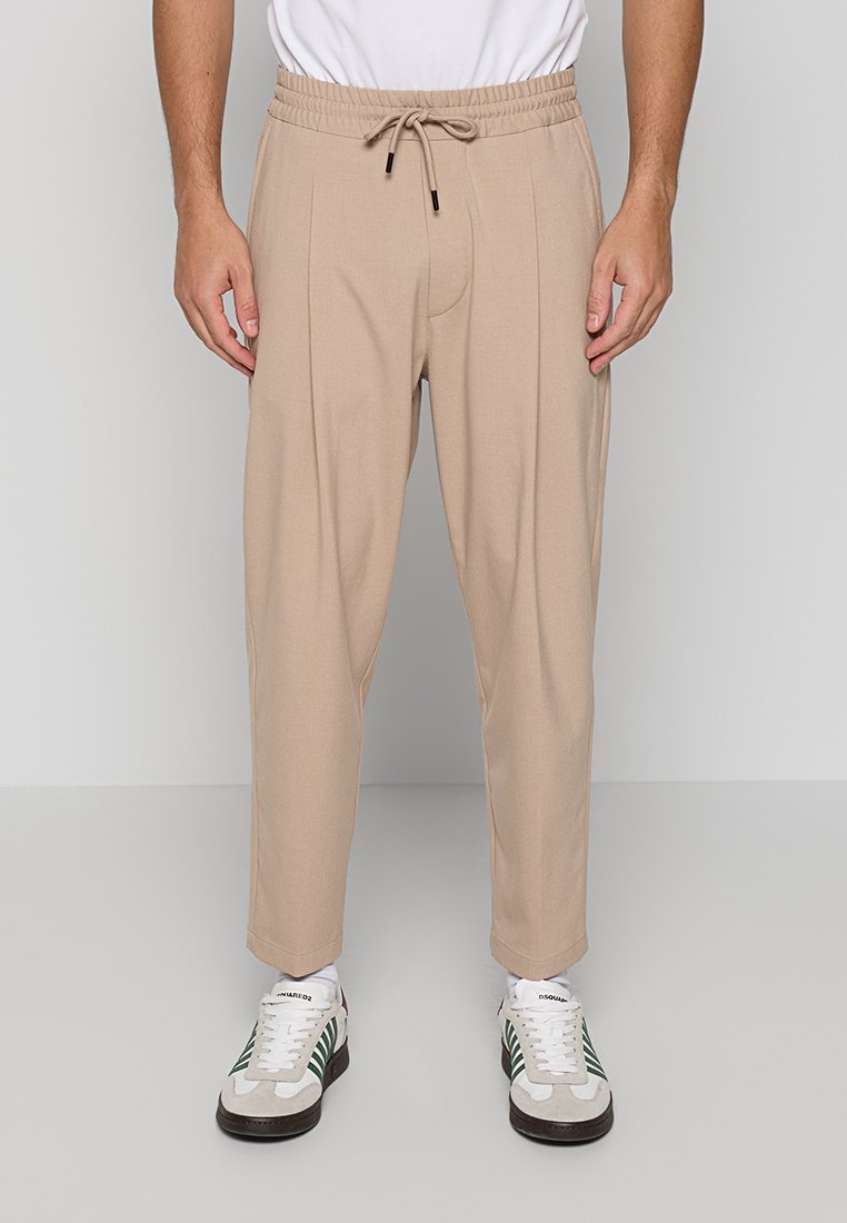 Mann trägt beige Kordelzughose und weiße Sneakers mit grünen Streifen, steht vor einfachem Hintergrund.