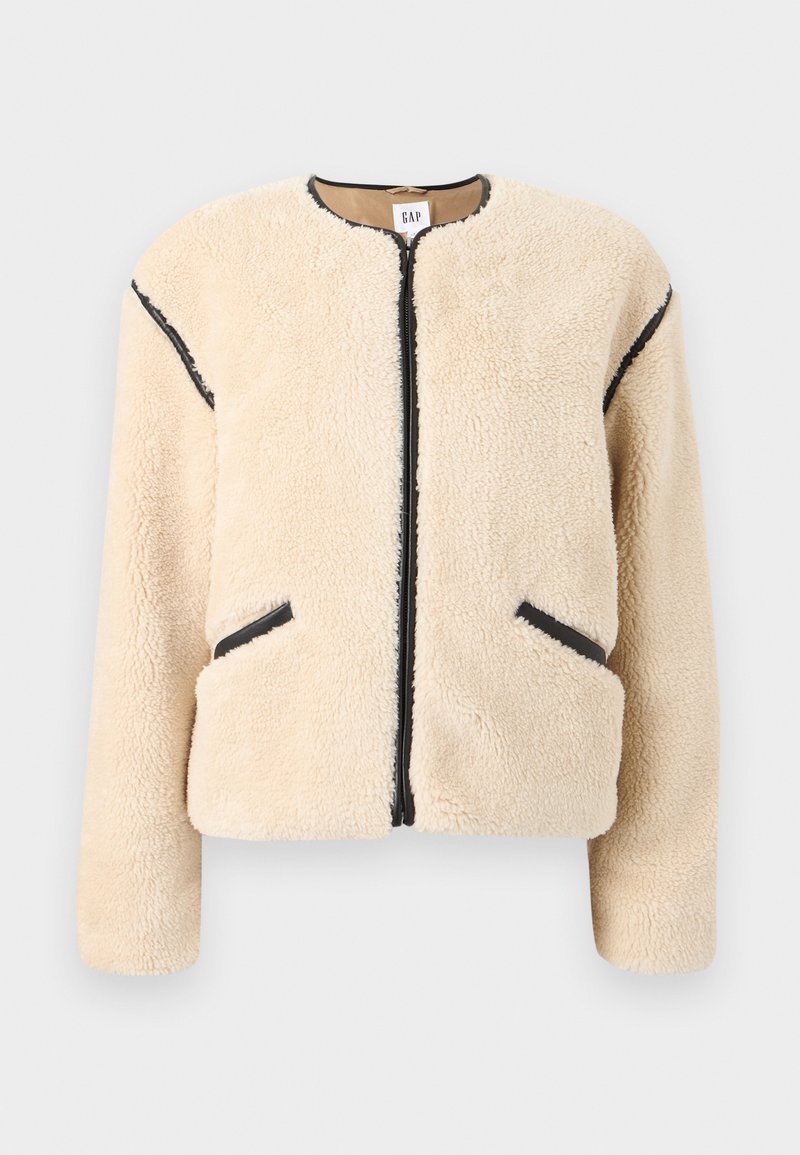 Beige Jacke aus Kunstpelz mit rundem Ausschnitt, Frontreißverschluss, schwarzen Akzenten an den Seiten und zwei Seitentaschen. Weiches, strukturiertes Material.