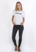 Frau steht barfuß mit Händen in den Taschen, trägt ein weißes Hurley-T-Shirt und schwarze enge Jeans vor einem weißen Hintergrund.