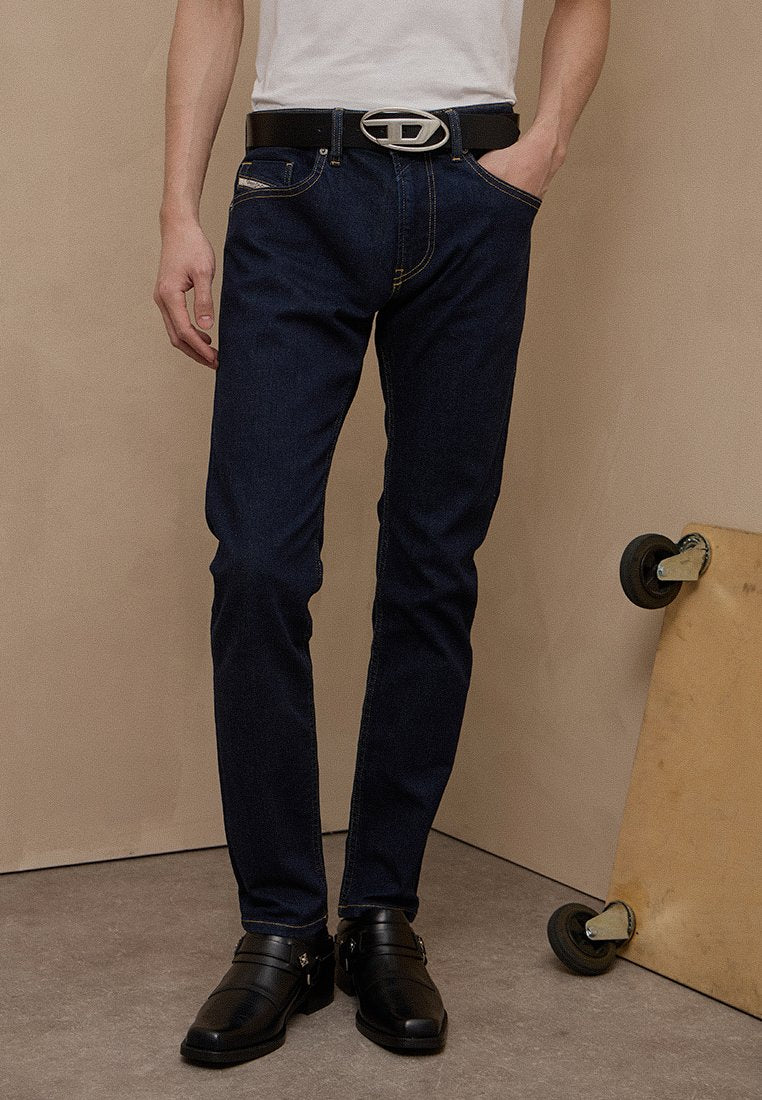 Mann trägt dunkelblaue Jeans mit schwarzem Gürtel und schwarzen Lederschuhen, steht neben einem an die Wand gelehnten Holz-Skateboard.