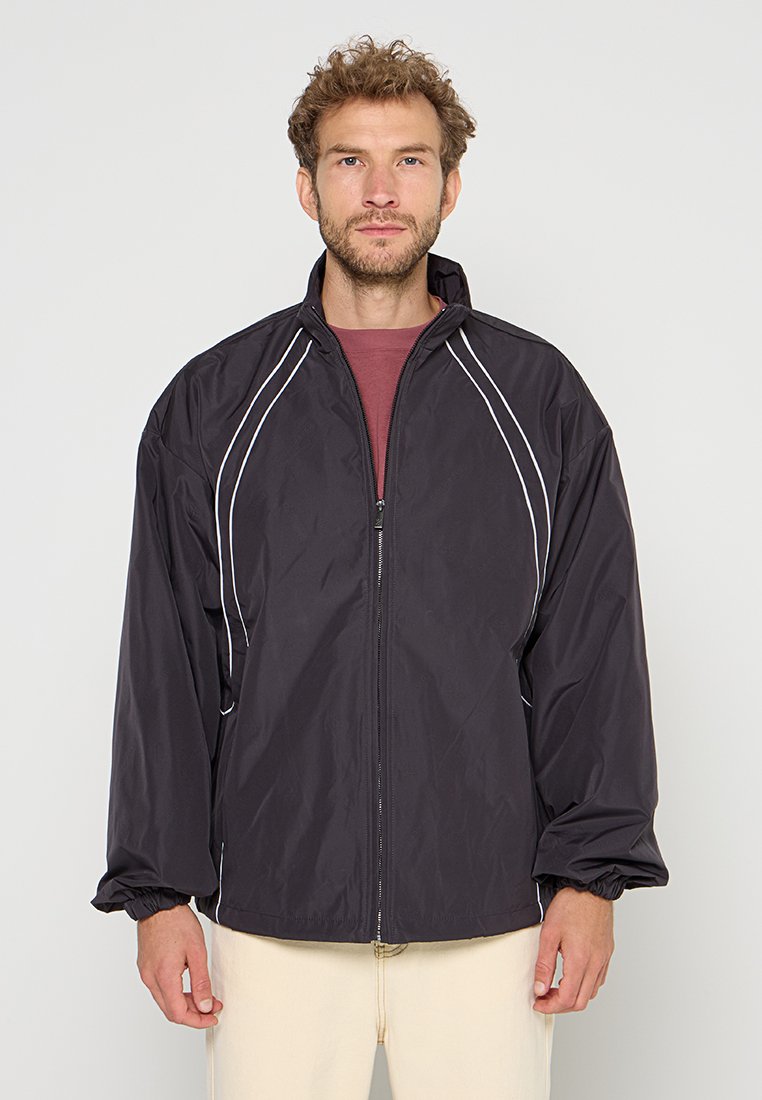 Mann mit lockigem Haar, der eine schwarze Zip-Jacke mit weißer Paspel über einem weinroten Shirt und hellbeigen Hosen trägt, steht vor einem schlichten Hintergrund.