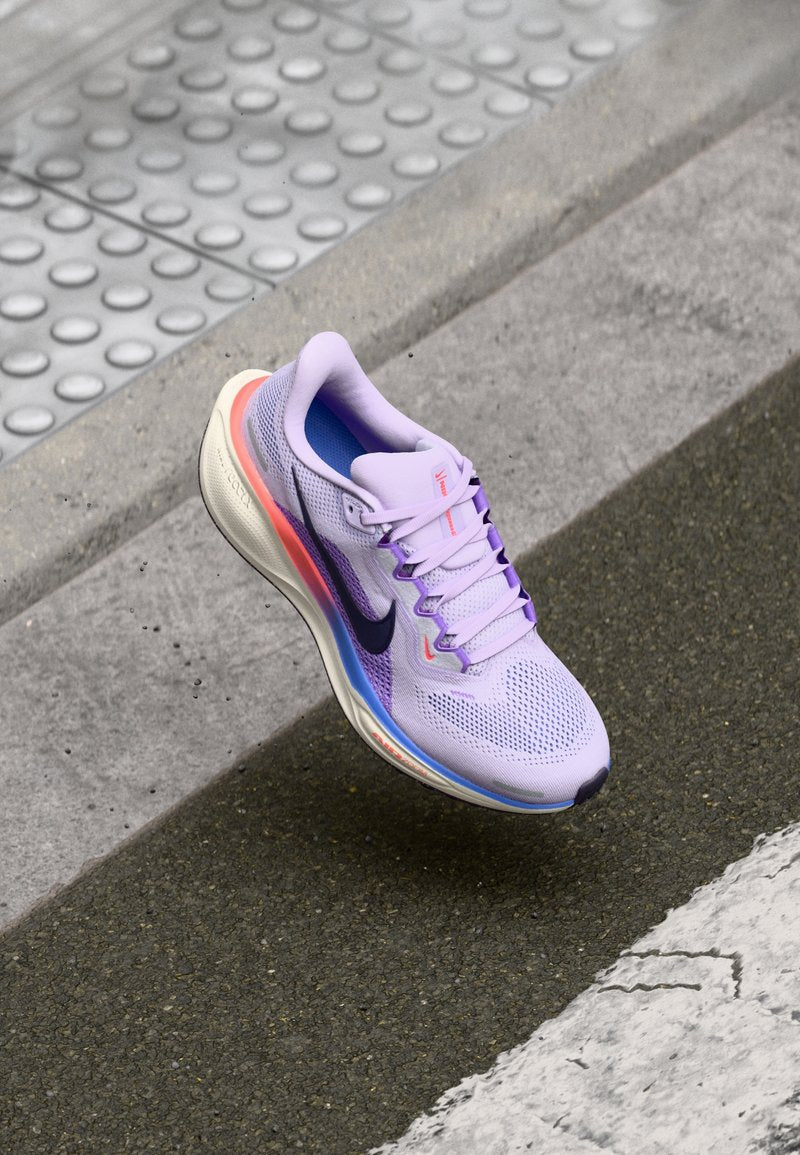 Lila Sportschuh mit atmungsaktivem Mesh-Obermaterial, kontrastierenden blauen und pinken Akzenten sowie einer gepolsterten Sohle, schwebend über einer strukturierten Oberfläche.