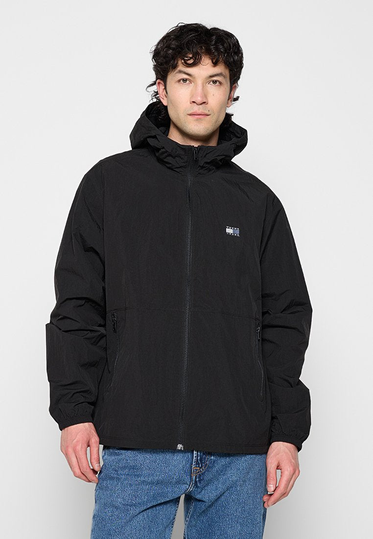 Mann trägt eine schwarze Kapuzenjacke mit Reißverschluss und elastischen Bündchen sowie blaue Jeans und steht vor weißem Hintergrund.