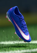 Blaue Fußballschuhe mit weißem Nike-Swoosh, rosa Akzenten und strukturiertem Finish. Über dem Gras erhöht, um ein dynamisches Design zu präsentieren.