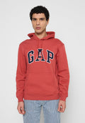 Mann mit lockigem Haar, der einen roten GAP-Kapuzensweater und blau-weiß gestreifte Hosen trägt, steht vor einem weißen Hintergrund.