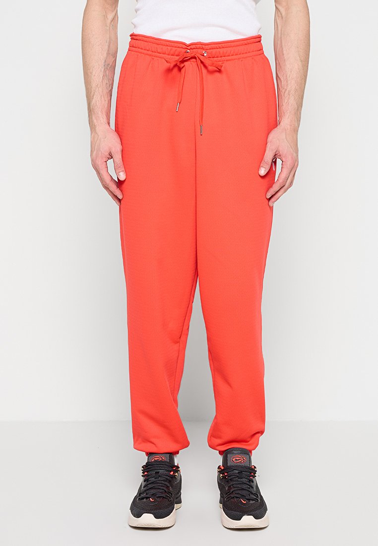 Mann trägt leuchtend orangefarbene Jogginghosen mit Kordelzug und schwarze Sportschuhe, steht vor weißem Hintergrund.