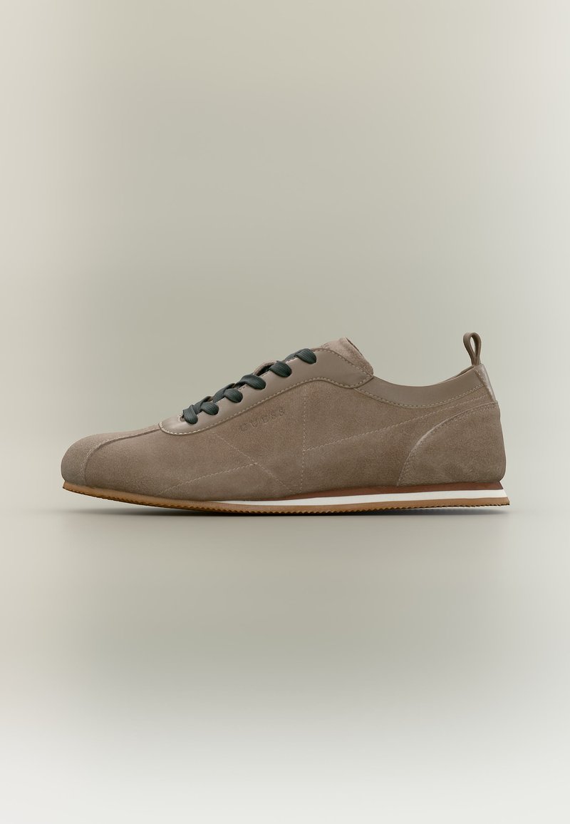 Hellbrauner Wildleder-Sneaker mit schwarzen Schnürsenkeln, abgerundeter Spitze, Zuglasche an der Ferse und flacher Gummisohle mit weißen und braunen Streifen.