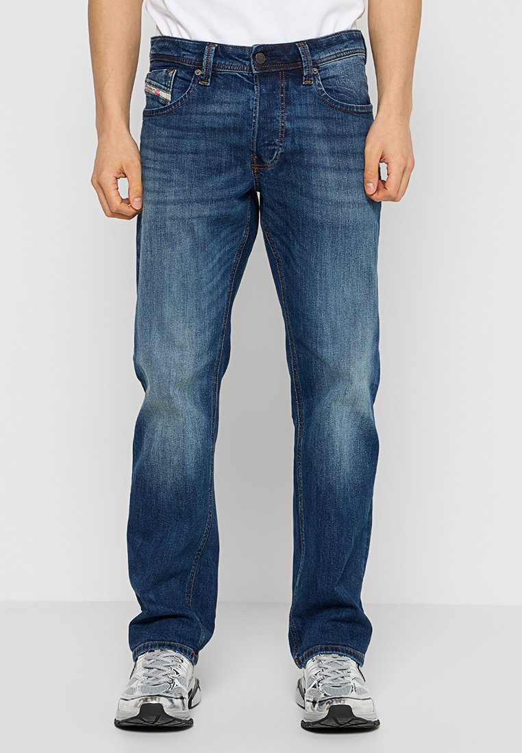 Dunkelblaue Jeans aus Denim mit geradem Schnitt, dezentem Fade-Effekt, fünf Taschen und einem Knopfverschluss. Getragen mit grauen Sneakers.