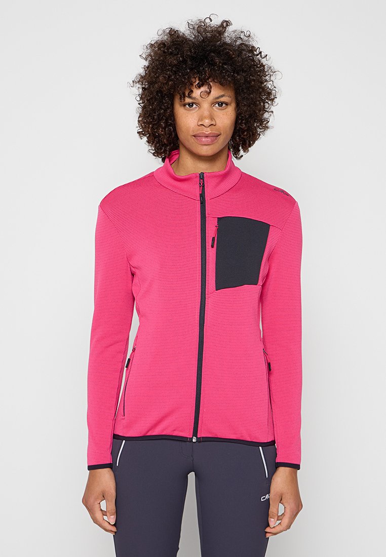 Frau mit lockigem Haar, die eine rosa Jacke mit Reißverschluss und einer schwarzen Brusttasche sowie dunkle Hosen trägt und vor einem einfarbigen Hintergrund steht.