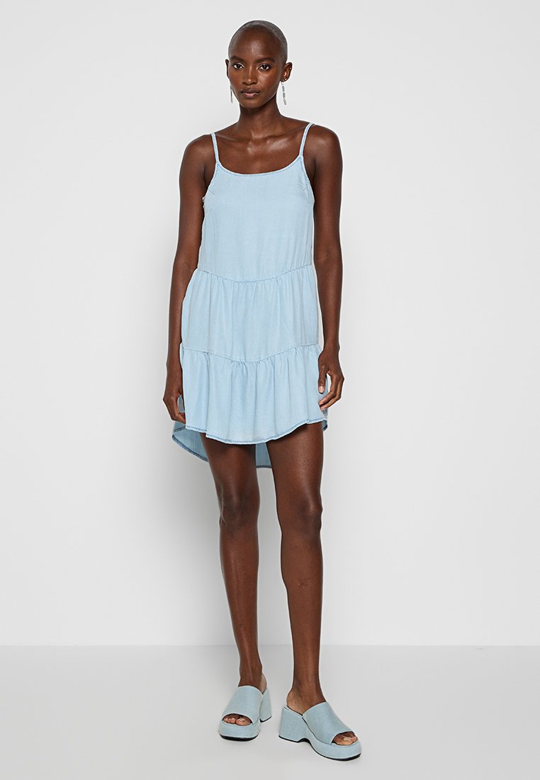 Hellblaues, ärmelloses Kleid mit einem gestuften Design und einem abgerundeten Saum. Kombiniert mit hellblauen Plateau-Schuhen. Glatte Stofftextur.
