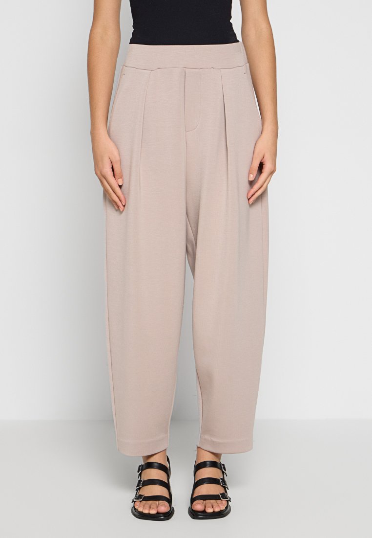Beige hoch taillierte Plisseehosen aus weichem Stoff, mit einem lockeren Schnitt und schmal zulaufenden Beinen sowie Seitentaschen, kombiniert mit schwarzen Riemchensandalen.